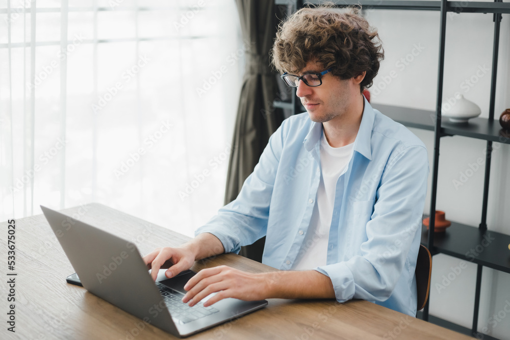 Happy young Caucasian man wearing glasses and smiling working typing on his laptop to get all his business done early in the morning with his cup of coffee. Young white man studying online at home. Happy young Caucasian man wearing glasses and smiling working typing on his laptop to get all his business done early in the morning with his cup of coffee. Young white man studying online at home.