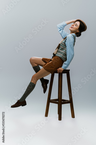 full length of positive young woman in beret and autumnal clothes sitting on wooden high chair on grey.