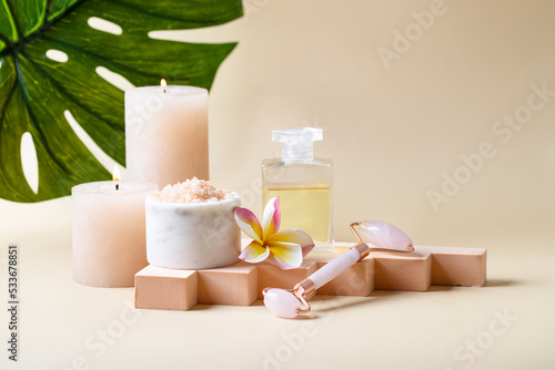 Beautiful SPA still life composition. Burning candles, bath salt, gua sha roller, natural massage oil, frangipani flower on the geometric podium on beige background with mosntera leaf. Copy space photo