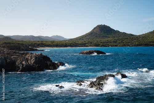The waves splashing on a rocky coastline of Cala Agulla (Cala Ratjada) on Mallorca in the mediterranean sea. Deep blue water and a green mountain (Puig de s’Àguila) in the background.
