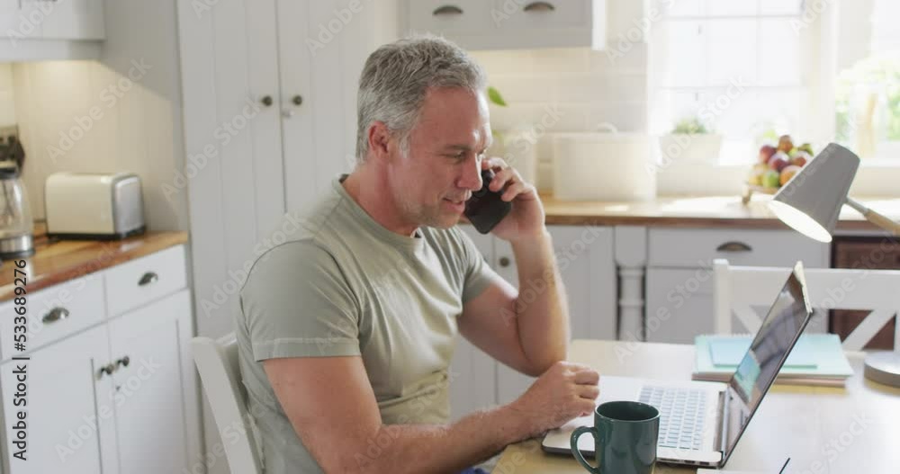 Happy caucasian man sitting at table in kitchen, using laptop and talking on smartphone Happy caucasian man sitting at table in kitchen, using laptop and talking on smartphone