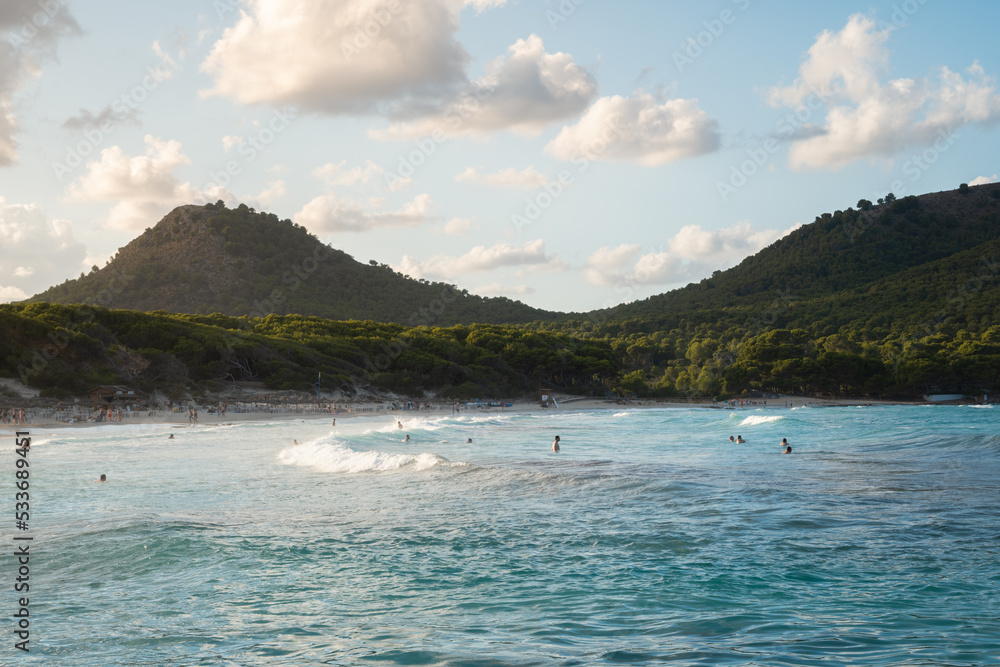 Obraz premium The waves splash on beach of Cala Agulla (Cala Ratjada) on Mallorca in the mediterranean sea. Blue water and a green mountain in the background. People bathing, surfers surfing. Tropical paradise.
