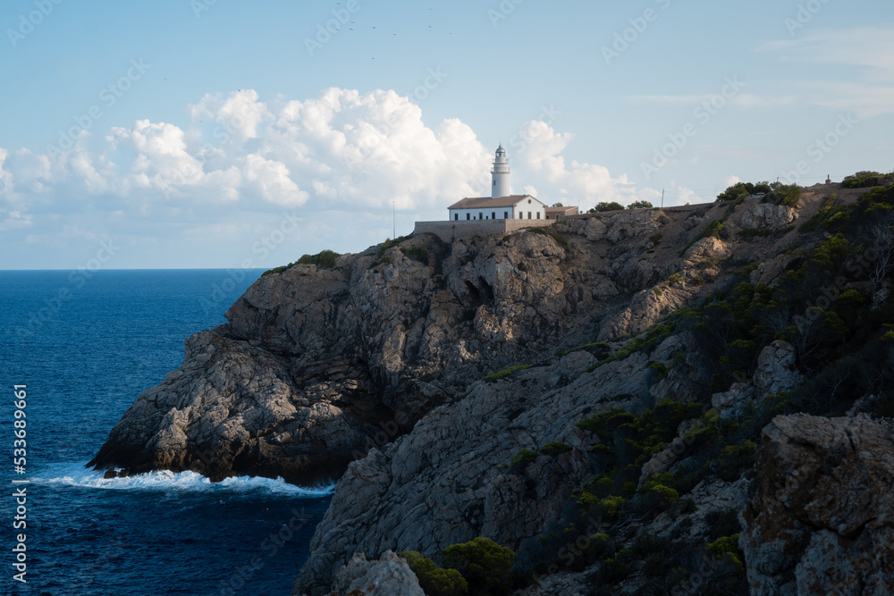 The lighthouse of Cala Ratjada. Waves splashing on a rocky coastline of Mallorca in the mediterranean sea. Deep blue water and steep cliffs. The lighthouse of Cala Ratjada. Waves splashing on a rocky coastline of Mallorca in the mediterranean sea. Deep blue water and steep cliffs.