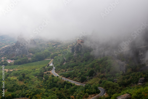 Scenic curvy mountain road with view of Holy Monastery of Rousanos appearing from the fog, Kalambaka, Meteora, Thessaly, Greece, Europe. Mystical landscape. Landmark build on dramatic rock formation