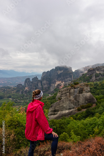 Woman with scenic view of Holy Monastery of Rousanos and Holy Monastery St Nicholas Anapafsas appearing from fog, Kalambaka, Meteora, Thessaly, Greece, Europe. Misty atmosphere in dramatic landscape