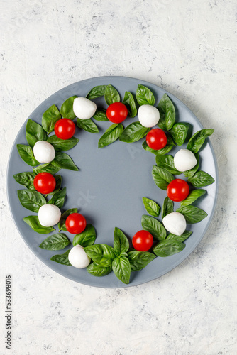 Caprese salad in the form of a Christmas wreath. Festive tomato mozzarella and basil appetizer on grey plate.