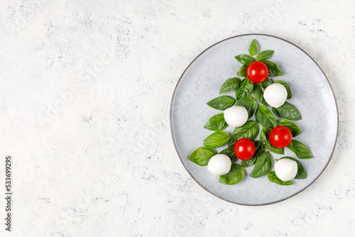 Caprese salad in the form of a Christmas tree. Festive tomato mozzarella and basil appetizer on light plate.
