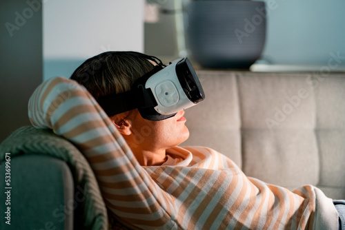 Happy woman lying on cozy sofa resting after hardworking day. Female wearing VR glasses plays interesting games with amazing graphics and explores virtual reality at home at night. Cyberspace concept