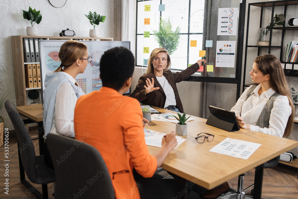 Senior blond financier woman pointing on board with paper sticks during speech on conference with multiracial female colleagues. Competent economists discussing company incomes at office room. Senior blond financier woman pointing on board with paper sticks during speech on conference with multiracial female colleagues. Competent economists discussing company incomes at office room.