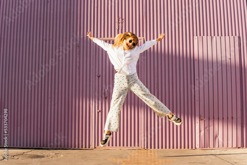 Cheerful woman jumping with arms outstretched in front of corrugated wall photo