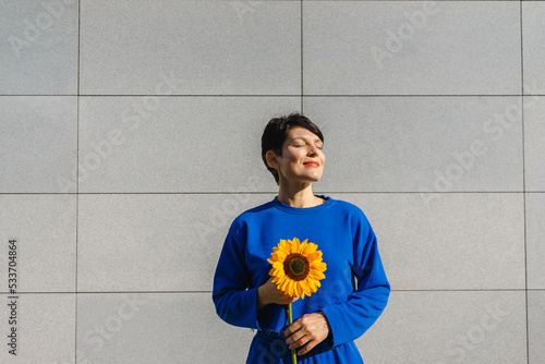 Smiling woman with sunflower enjoying sunlight in front of wall photo