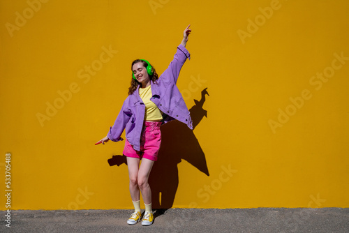 Carefree woman with hand raised dancing and listening to music in front of yellow wall photo