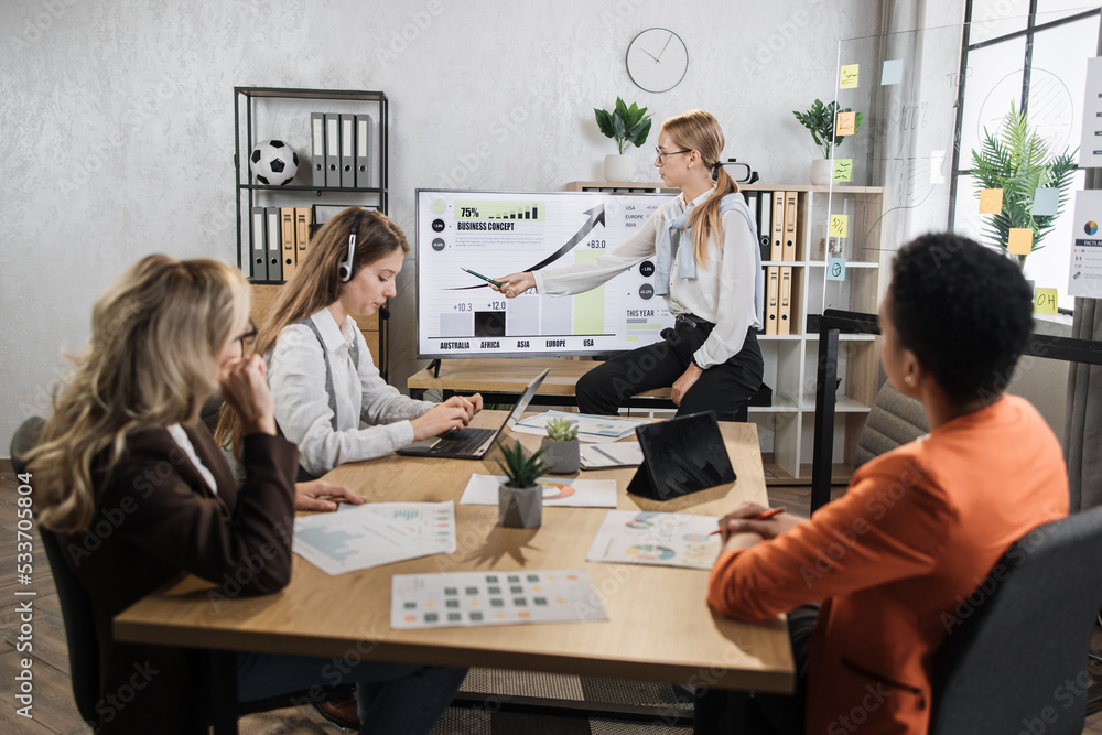 © sofiko14 - Caucasian woman pointing on monitor with financial report during conference with multiracial female partners. Business woman colleagues gathering at office room for brainstorming.