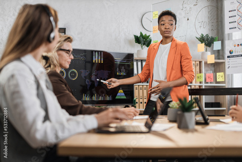 African american woman in orange suit showing financial statistic of enterprise during working meeting with diverse female colleagues. Cooperation  technology and people concept.