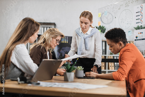 Young businesswoman in white shirt smiling and showing financial report during conference with multiracial partners. Business people sitting at desk office with papers and modern technology.