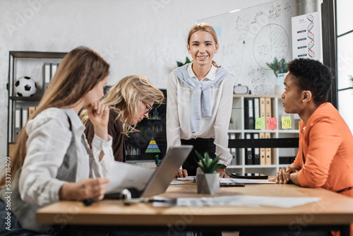 Multi ethnic company workers sitting at desk with modern devices, preparing financial report and listening speech of their young female boss. Concept of technology and cooperation.