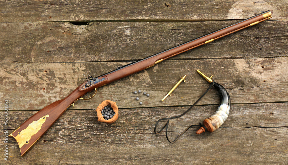Traditional Kentucky rifle and shooting equipment such as a gunpowder horn,  a gunpowder measure and bullets on an old, damaged board. Stock Photo