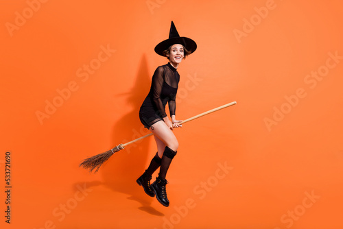 Full body photo of nice young woman straddle wooden broom fly wear stylish black halloween witch look isolated on orange color background photo