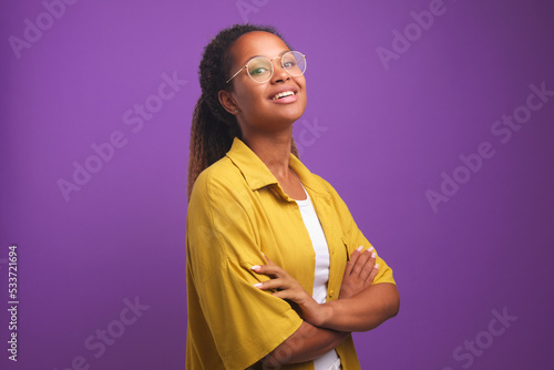 Young confident positive African American woman proudly posing with arms crossed in front of chest and raising chin looks at screen dressed in yellow shirt stands on purple studio background