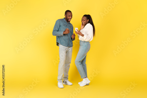 Excited African American Couple Using Mobile Phone Over Yellow Background