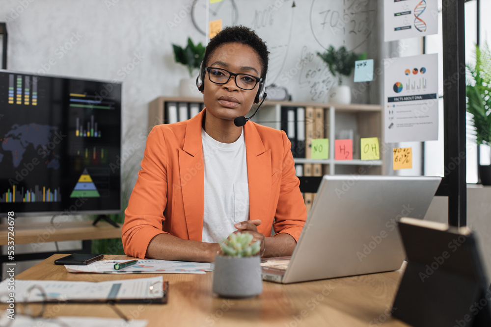 Portrait of attractive african business woman in headset sitting at office desk and looking at camera. Business lady having video conference. Business meeting online. Portrait of attractive african business woman in headset sitting at office desk and looking at camera. Business lady having video conference. Business meeting online.
