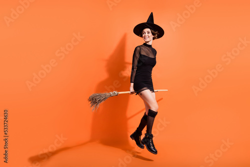 Full body photo of cute girl sit flying broom look copyspace dressed trendy black halloween garment isolated on orange color background photo