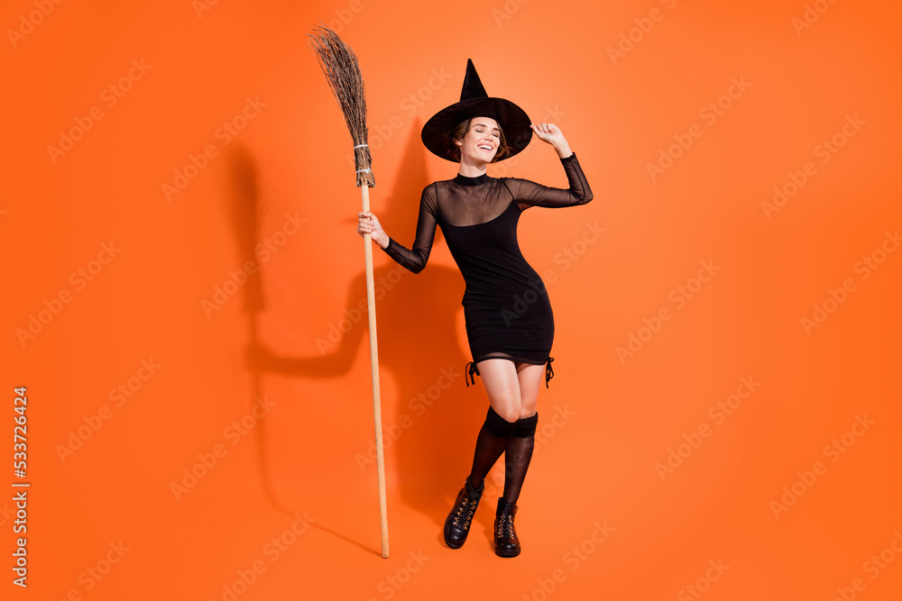 Full body photo of pretty young girl touch hat hold broom dressed trendy black halloween witch clothes isolated on orange color background Full body photo of pretty young girl touch hat hold broom dressed trendy black halloween witch clothes isolated on orange color background