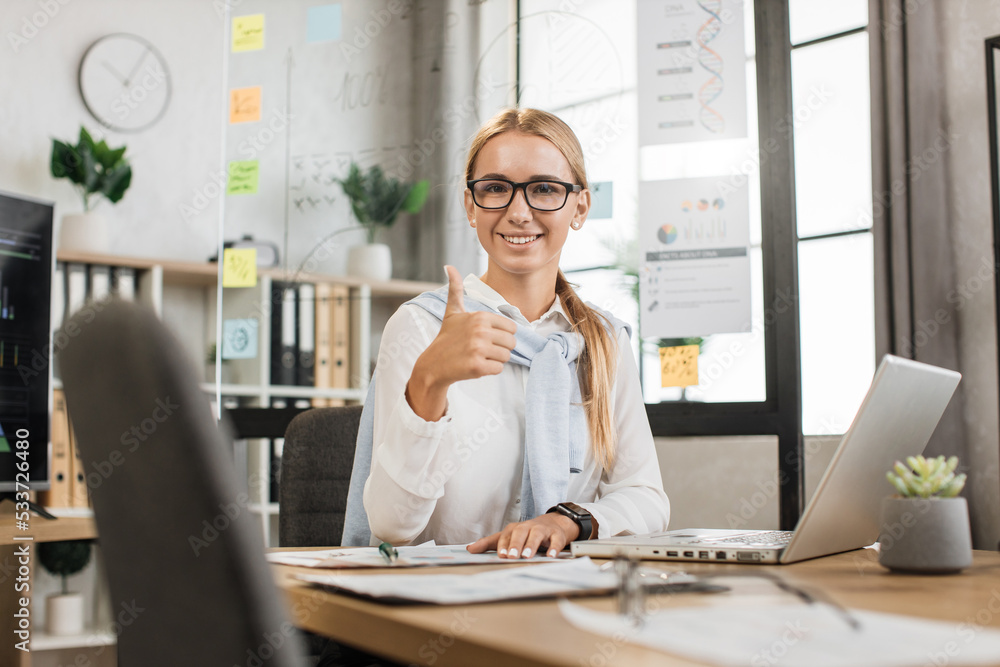 Busy caucasian woman in white shirt sitting at office and using wireless laptop showing thumb up. Big monitor with various charts and graphs on background. Business and finance concept. Busy caucasian woman in white shirt sitting at office and using wireless laptop showing thumb up. Big monitor with various charts and graphs on background. Business and finance concept.
