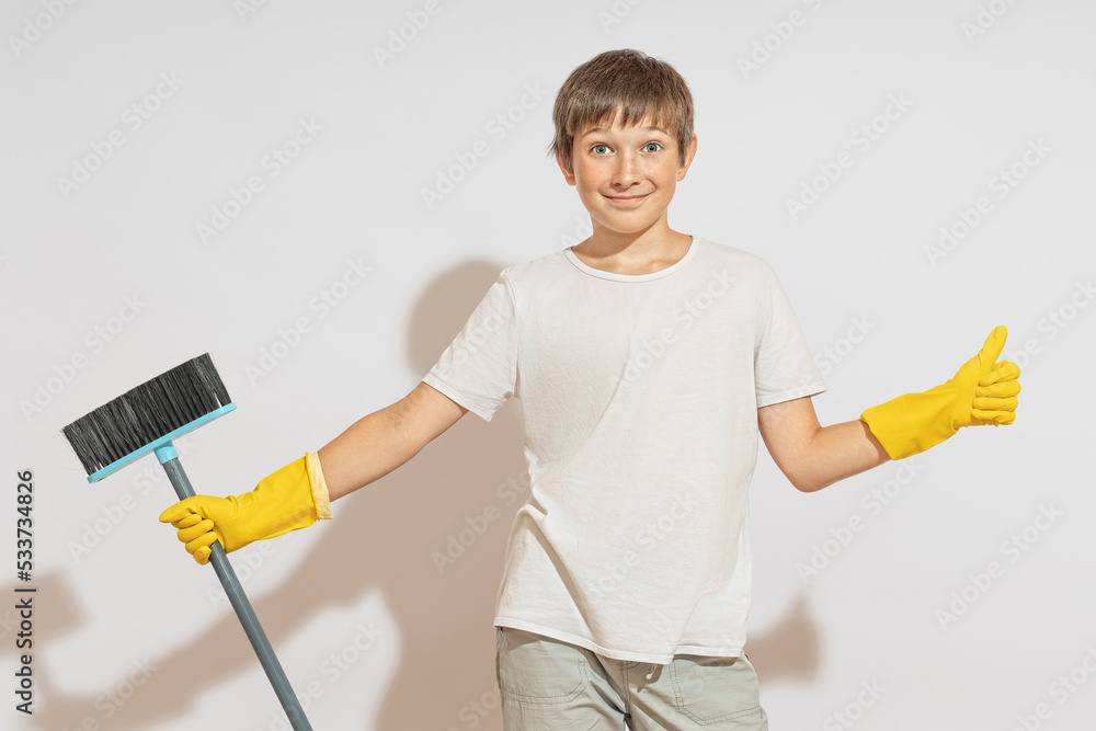 A teenager in rubber gloves smiles at the camera, shows a like sign and holds a floor brush. The concept of readiness for cleaning.
