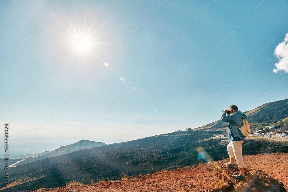 Rear view of man looking through binoculars at panoramic view of colorful summits of active volcano Etna, Tallest volcano in Continental Europe, Sicily, Italy.