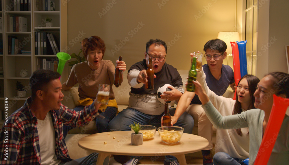 Obraz premium Happy asian friends or football fans watching soccer on tv and celebrating victory at home.Friendship, sports and entertainment concept.