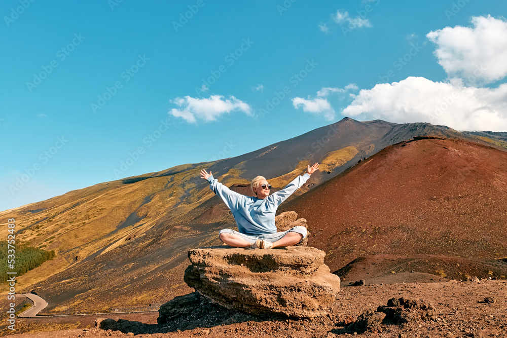 Happy tourist woman enjoying freedom while doing yoga on big stone at panoramic view of colorful summits of active volcano Etna, Tallest volcano in Continental Europe, Sicily, Italy.