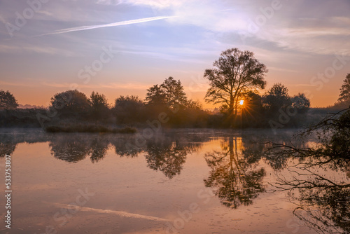 Krajobraz jesienny. Mglisty jesienny poranek nad dziką rzeką. Poranna mgła nad wodą. Brzask i promienie słońca nad rzeką. Odbicia w wodzie. © anettastar