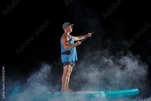 A man on a sub board with an oar in his hands on a black background in the fog. horizontal photo