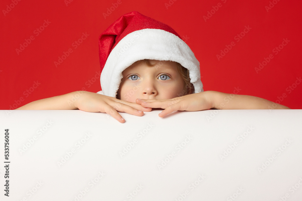 Serious little boy stares intently at camera with his hands folded on white table. Sad child in Christmas red hat on red background folded his hands on banner Serious little boy stares intently at camera with his hands folded on white table. Sad child in Christmas red hat on red background folded his hands on banner