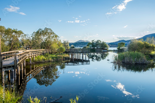 A wood bridge wraps along the edge of a pond reflecting in the calm waters in Kelowna British Columbia.