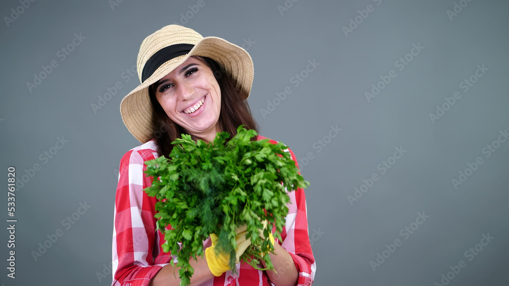 portrait of smiling female farmer in plaid shirt, gloves and hat holding, smells a bunch of fresh green parsley, greens, on gray background, in studio. Healthy food to your table. High quality photo