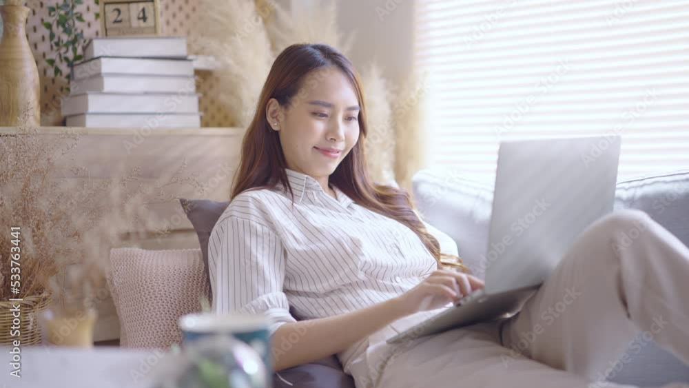 Young Asian woman using laptop on sofa living room at home cozy happy working at home. Young Asian woman using laptop on sofa living room at home cozy happy working at home.