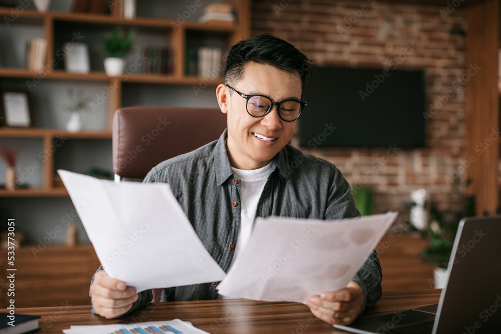 Smiling adult korean male in glasses works with papers, charts and graphs at table in office interior