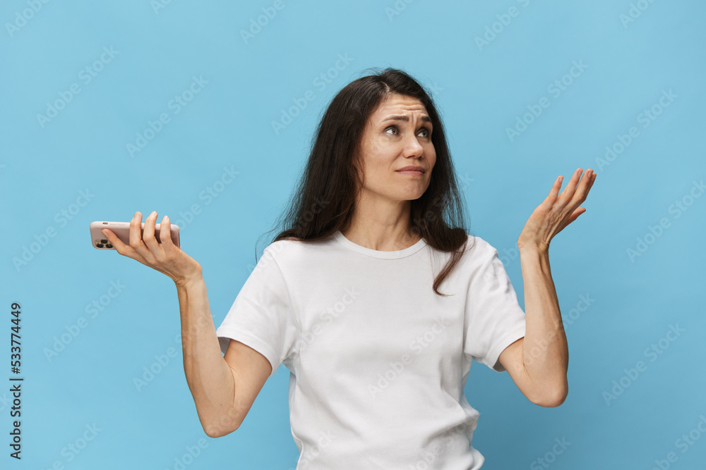 Obraz premium a pensive woman spreading her arms in a white T-shirt stands on a blue background with a phone in her hands and looks at the top of the background