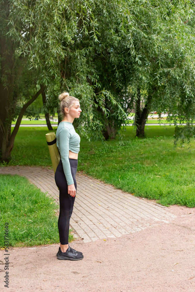 A slender woman in sportswear, standing in summer in park, with a green gym mat for sports.