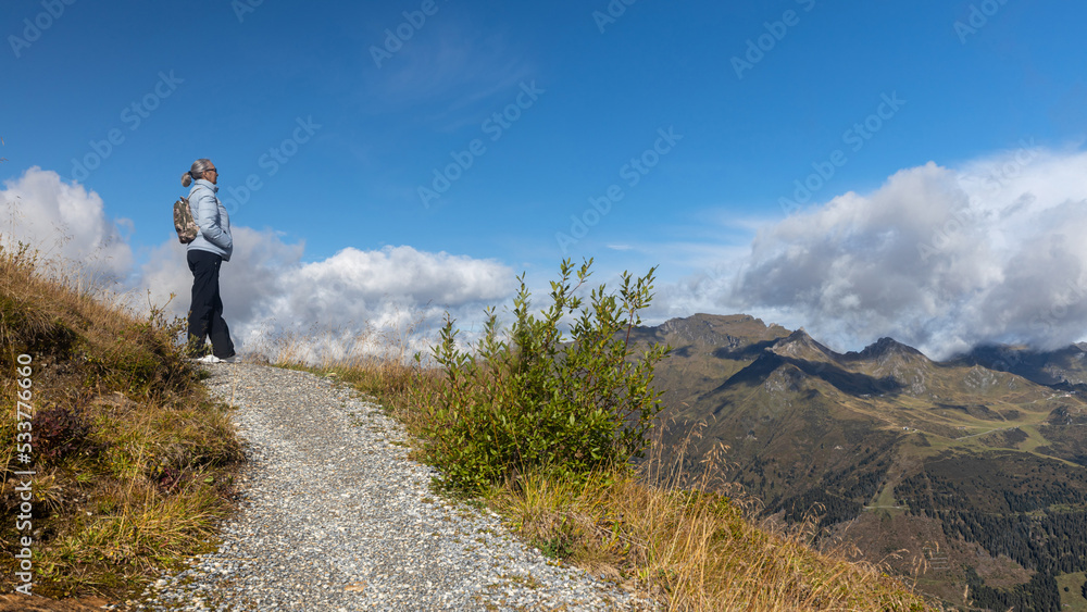 A gray-haired woman stands on a mountain road and looks at a beautiful landscape in the Austrian Alps