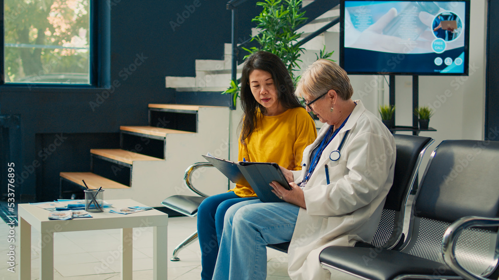 Elderly doctor and patient doing checkup visit in facility lobby, talking about treatment and medication. Taking notes on tablet and filling in medical report form to receive medical insurance. - obrazy, fototapety, plakaty 