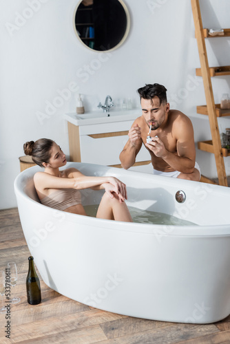 shirtless muscular man lighting cigarette near sexy girlfriend relaxing in bathtub.