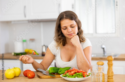Young housewoman eating vegetable salad from plate at home kitchen