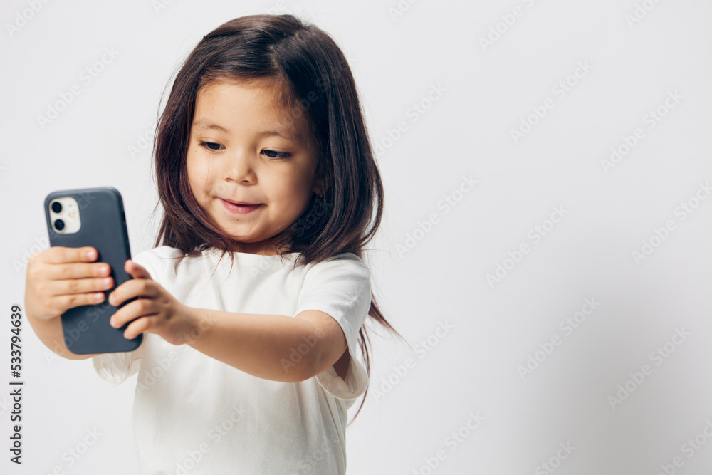 a cute little preschool girl stands on a white background in a white T-shirt and takes pictures of herself on her phone, smiling happily at the camera. The theme of children's happiness