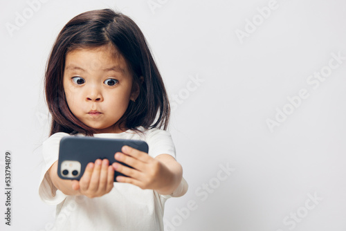 cute, funny, little girl in a white t-shirt stands on a light background with a smartphone in her hand and takes a selfie. Horizontal studio photography with blank space for advertising mockup insert