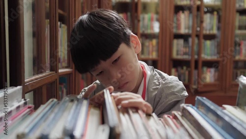 Cute and smart looking Asian elementary student child looking and searching for book from wooden bookshelf in library at school. Self study, reading lifestyle and back to school concept.