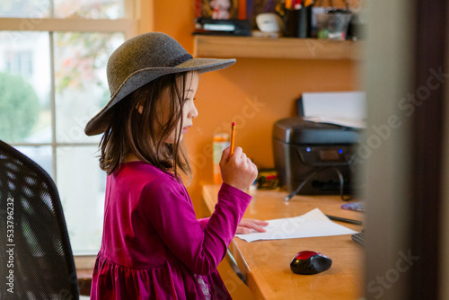 A small child in hat sits at desk with paper and pencil photo