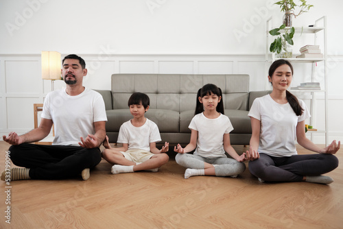 Happy Asian Thai family, parents, and children sit on living room floor, close their eyes together, tranquil meditate in lotus position, and practice healthy yoga, domestic home lifestyle weekend.